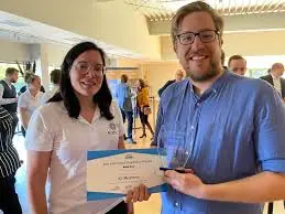 A woman and a man in white and blue shirts holding an award certificate at the Belgian Awards 2021