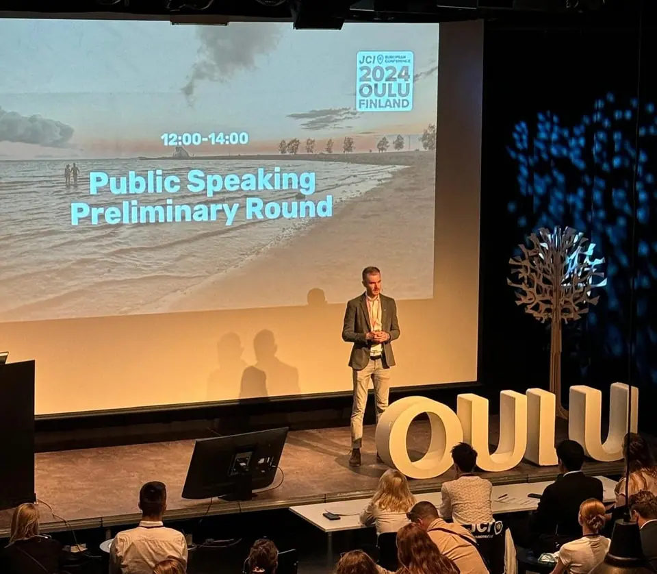 A speaker on stage in front of a Public Speaking Preliminary Round slide and a large OULU sign
