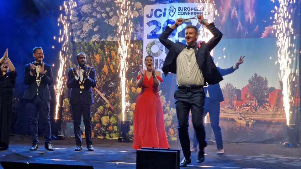 A man with raised arms and two award presenters on stage with pyrotechnic sparks at the gala