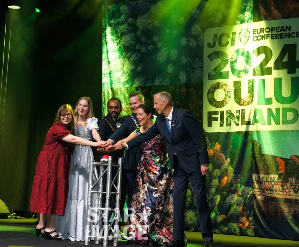 Six people in evening wear pressing a button together on stage in front of a green 2024 JCI European Conference Oulu backdrop