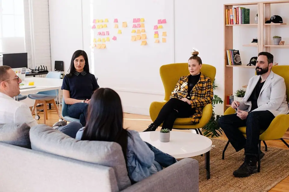 Four people in conversation on sofas and armchairs in a bright office, with a wall of sticky notes behind them