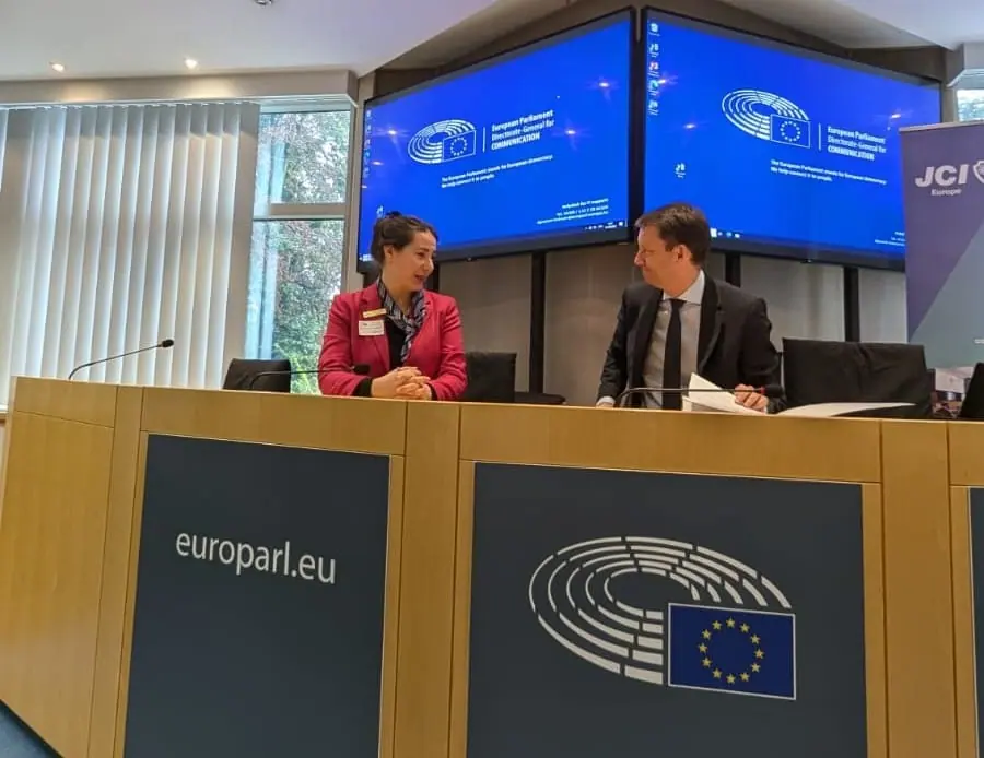 A woman and a man seated at a desk in a European Parliament meeting room with europarl.eu signs