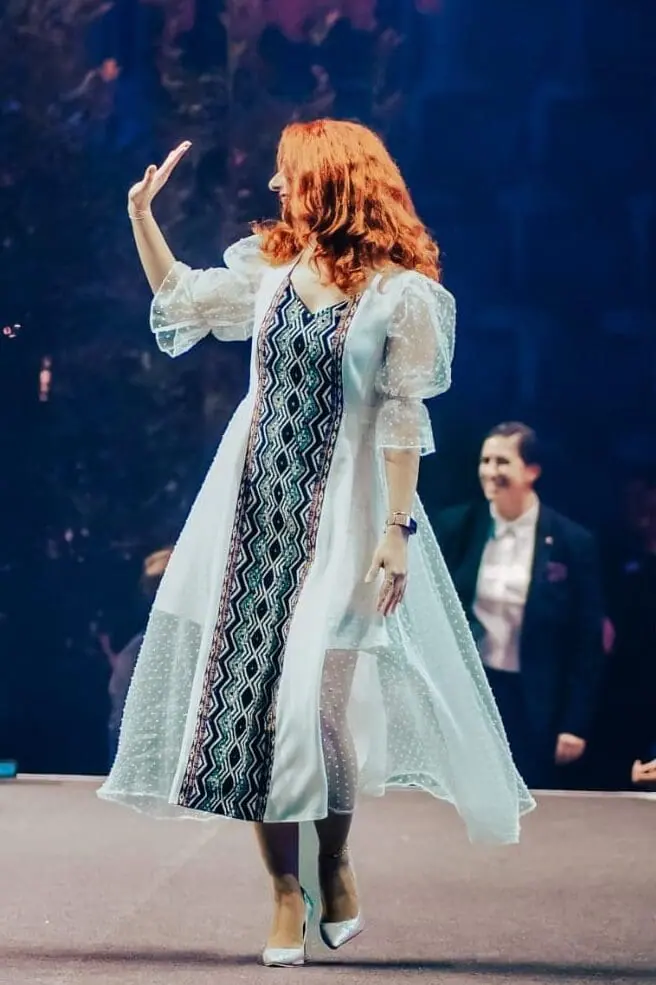 A woman in an embroidered white dress waving on a stage