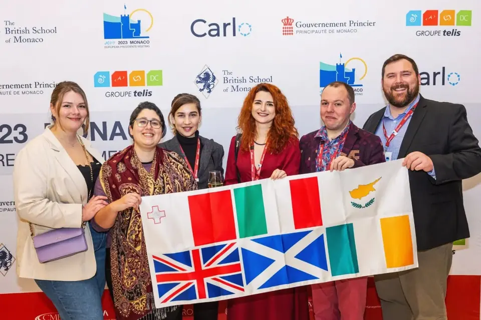 Six JCI members holding a banner with the flags of Malta, Italy, Cyprus, Ireland and the United Kingdom, in front of a Monaco sponsor wall