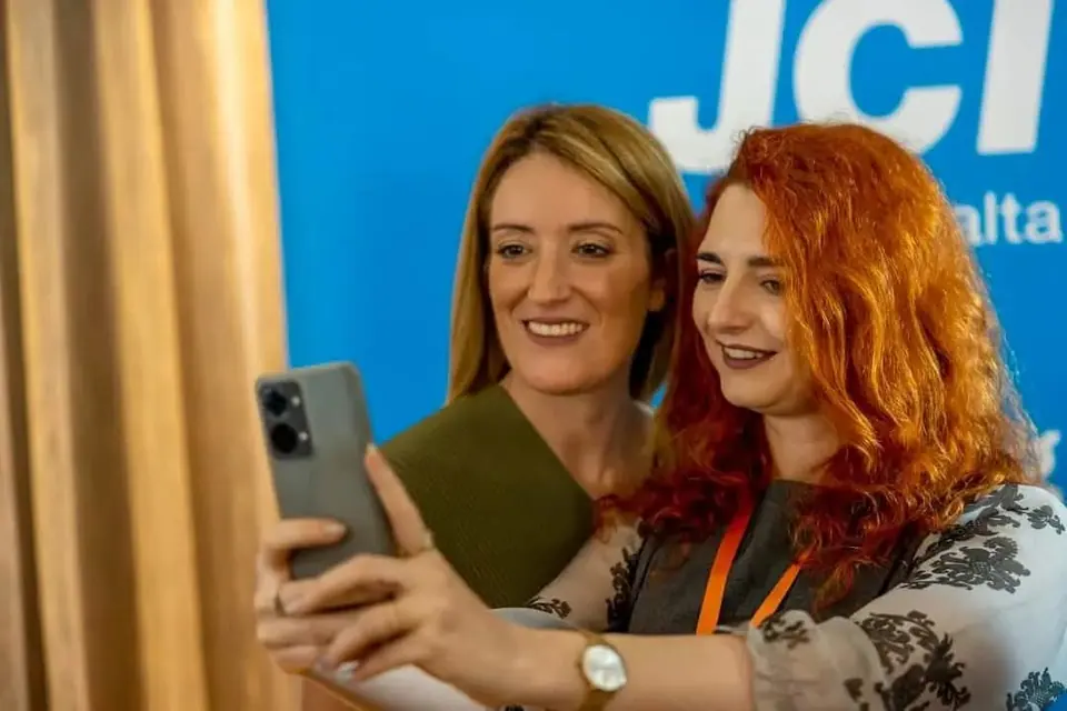 Alexandra Botoș taking a selfie with another woman in front of a JCI Malta banner