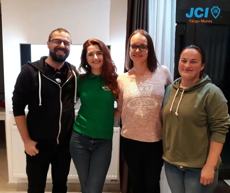 Four people in casual clothes posing in front of a JCI Târgu Mureș banner