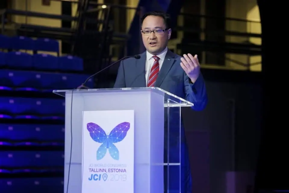 Kevin Hin speaking at a glass lectern with a butterfly logo for the Tallinn, Estonia World Congress