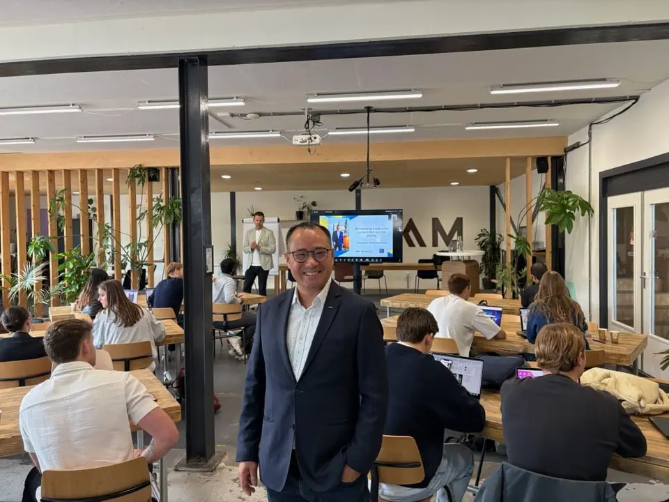 A man in a suit standing in a modern lecture hall with students working at tables