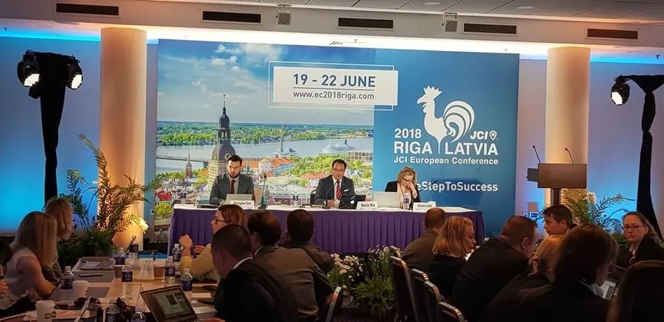 Officers at the head table in front of a Riga 2018 backdrop, with delegates seated in front