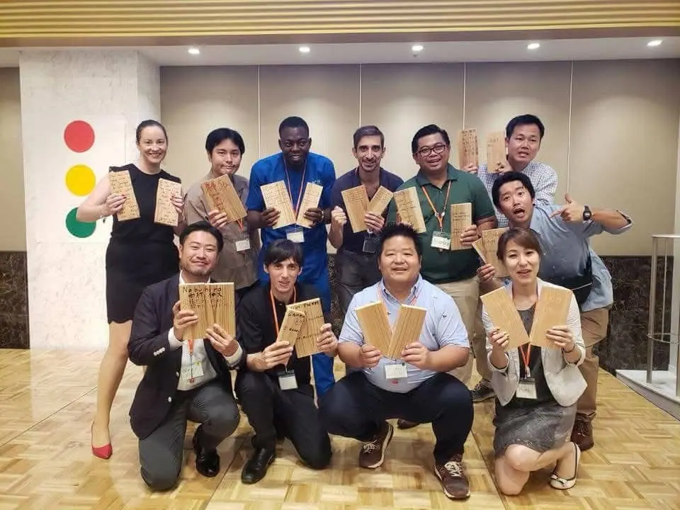 Thirteen participants kneeling and standing, holding small wooden boxes, in a hotel hall