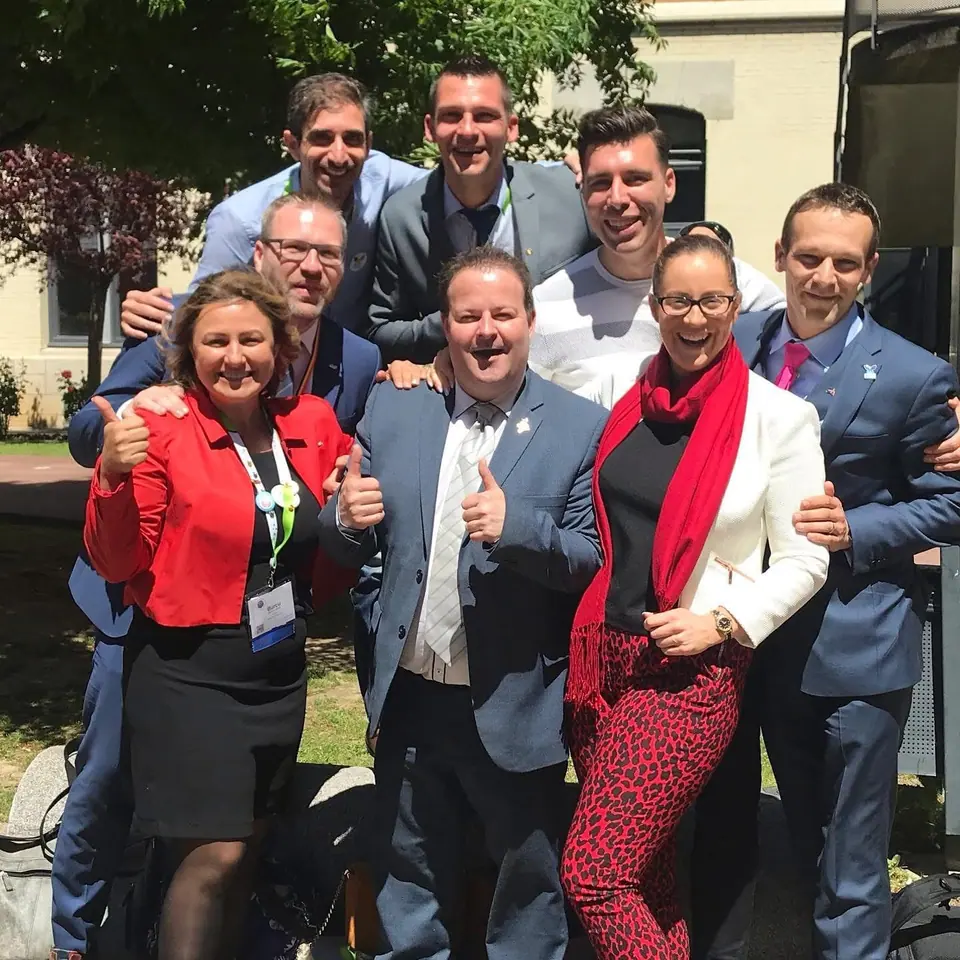 Seven smiling men and women giving thumbs up outdoors