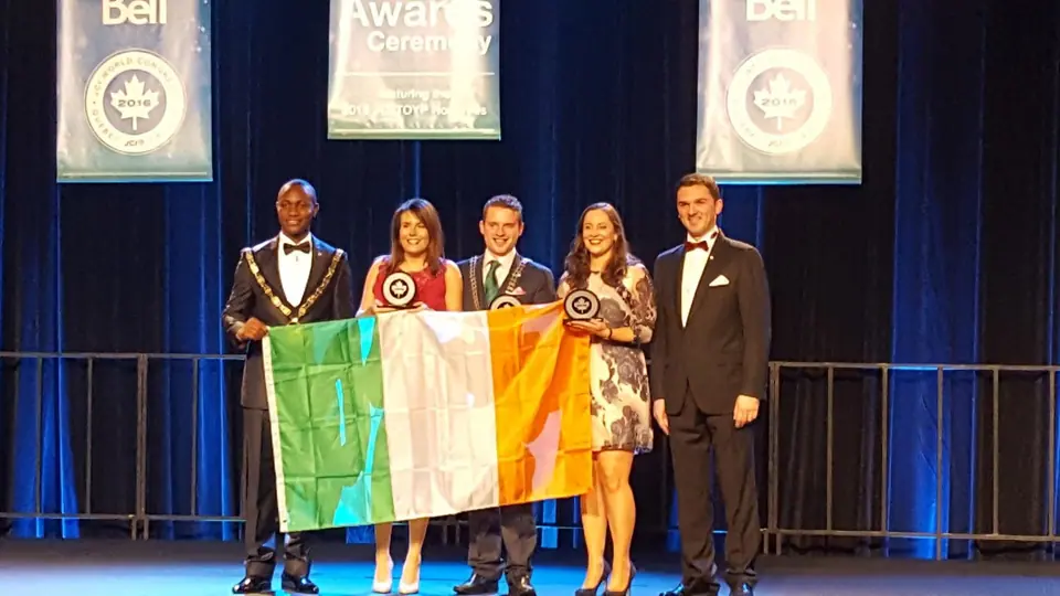 Two men in tuxedos, two women, and a woman in a gown holding Irish flags on a stage with award banners