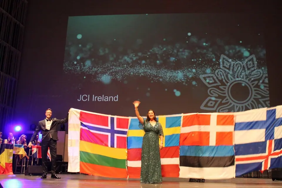 A man in a tuxedo and a woman in a green gown waving in front of a row of Nordic and Baltic flags on stage, under a JCI Ireland slide