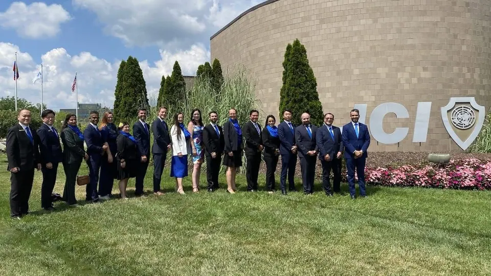 A group of about twenty JCI members in dark suits standing in front of a large JCI sign