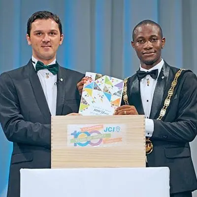 Two men in tuxedos, one wearing a chain of office, holding a box marked JCI 100