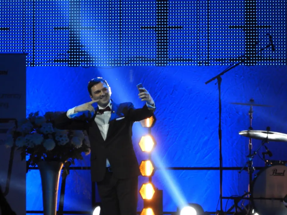 A man in a tuxedo taking a photo of the audience from a stage lit in blue