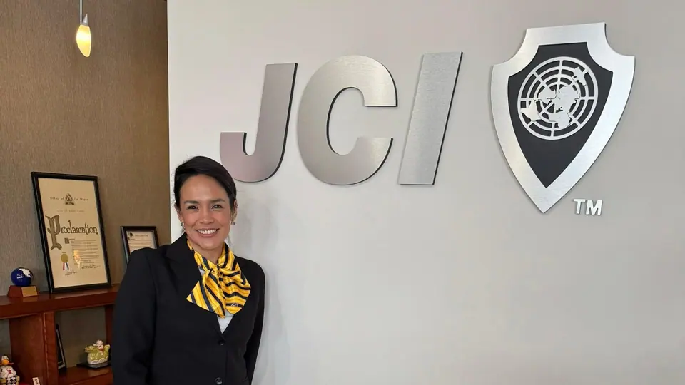 Alejandra Castillo in a dark jacket and yellow scarf standing in front of a large JCI logo