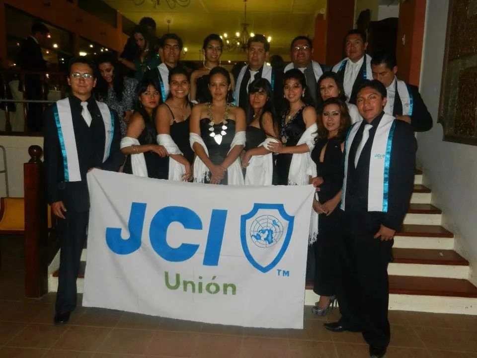 A group in dark clothes with sashes holding a JCI Unión flag on a staircase
