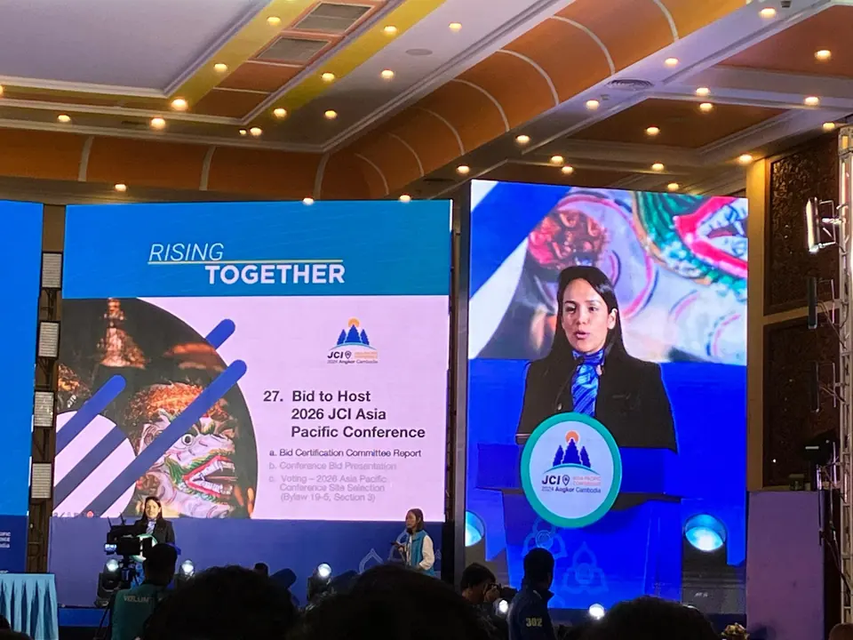 A speaker on a conference stage with a slide about the bid to host the 2026 JCI Asia-Pacific Conference