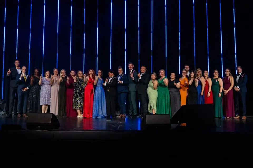 A line of people in formal wear on a dark stage giving thumbs up
