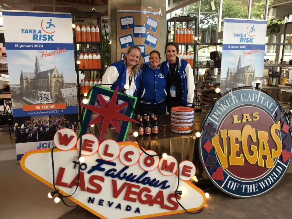 Three people in JCI jackets behind a Welcome to Las Vegas sign and Dare to Risk banners