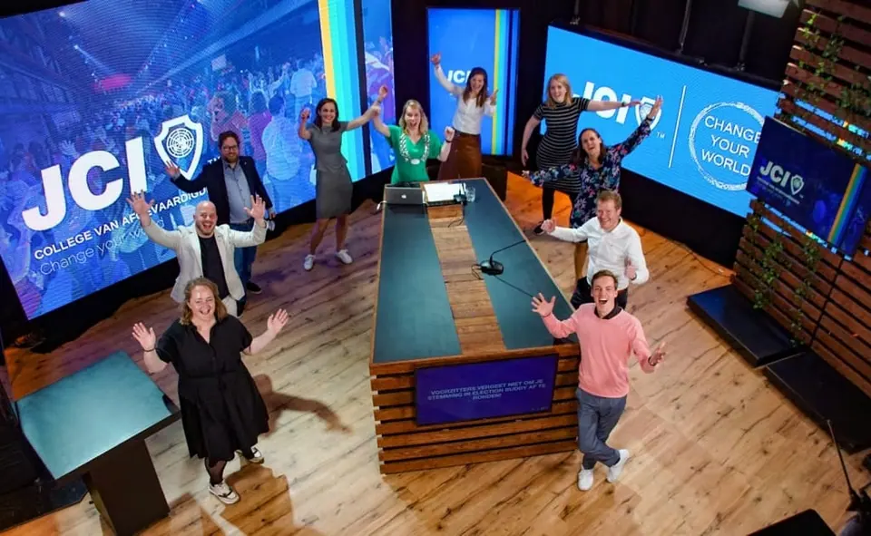 A JCI TV-style studio with people waving from the floor under large screens