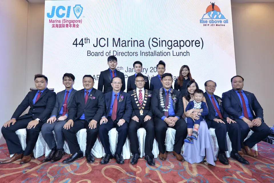 A board of directors seated in two rows in front of a JCI Marina (Singapore) installation lunch backdrop