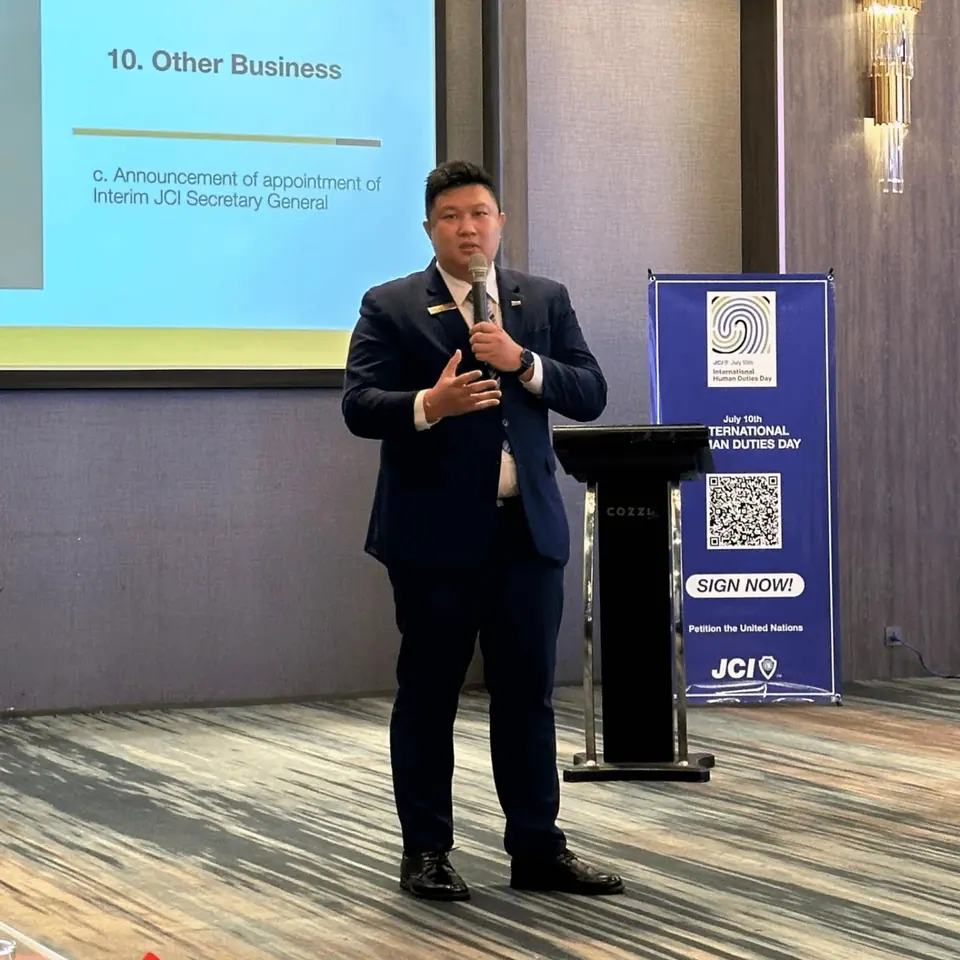 A man in a dark suit speaking in front of a presentation slide and a JCI banner