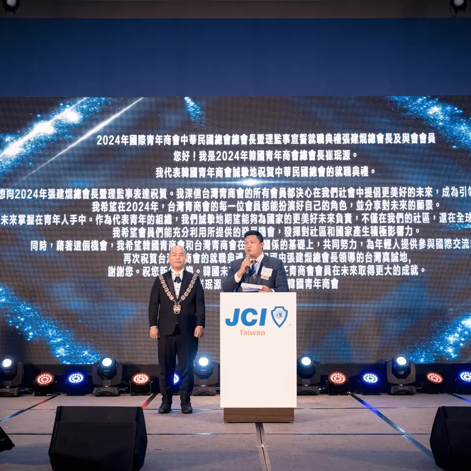 Two people on a stage, one speaking at a lectern, in front of a screen with Chinese text