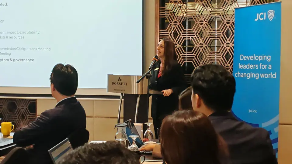 A woman speaking at a lectern in a seminar room next to a JCI banner reading Developing leaders for a changing world