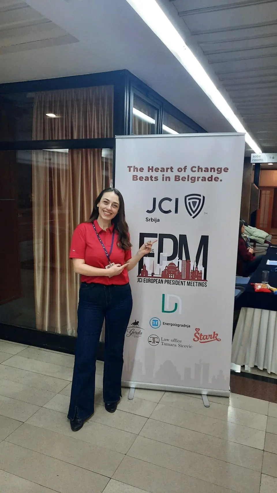 A woman in a red top standing next to a JCI banner reading The heart of change beats in Belgrade