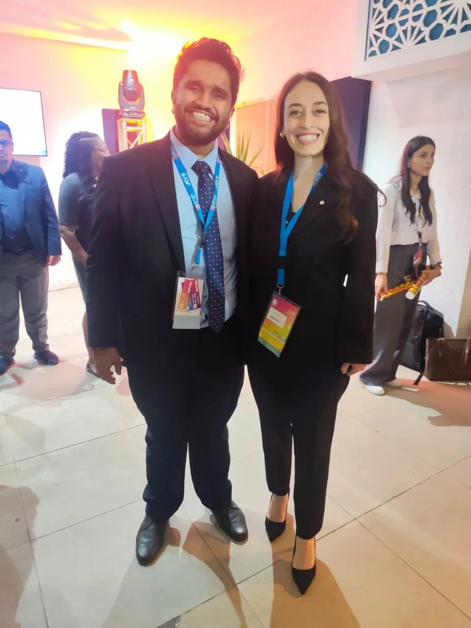 A man and a woman in dark suits with conference badges smiling at the camera