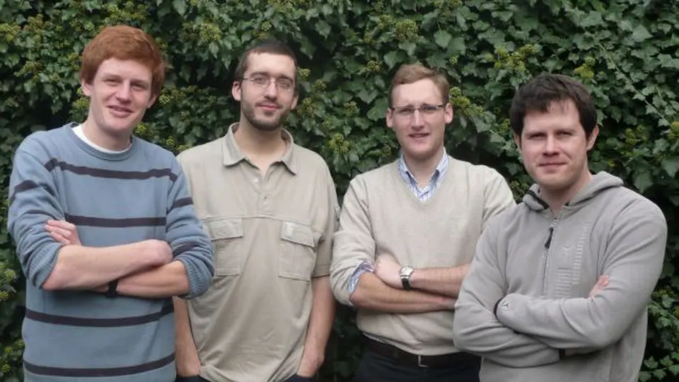Four young men in casual sweaters posing in front of an ivy hedge