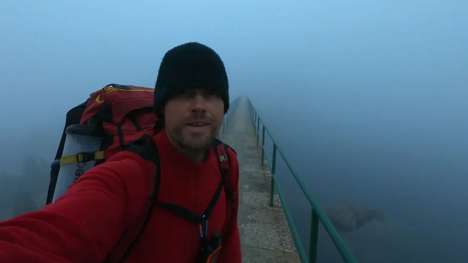 A hiker in a beanie and red jacket with a backpack taking a selfie on a narrow walkway in fog