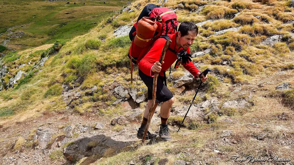 A hiker in a red jacket with trekking poles climbing a steep grassy mountainside