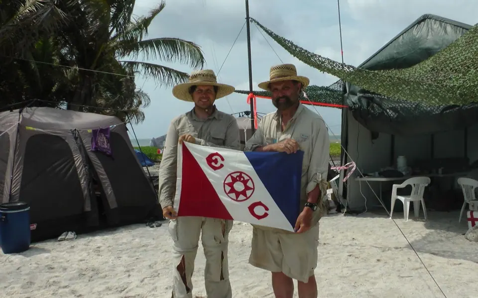 Two men in explorer hats holding the Explorers Club flag in front of tents on a beach