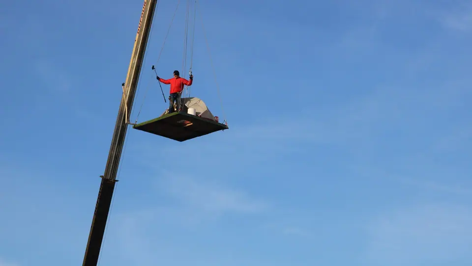 A person standing on a small platform hanging from a crane against a blue sky