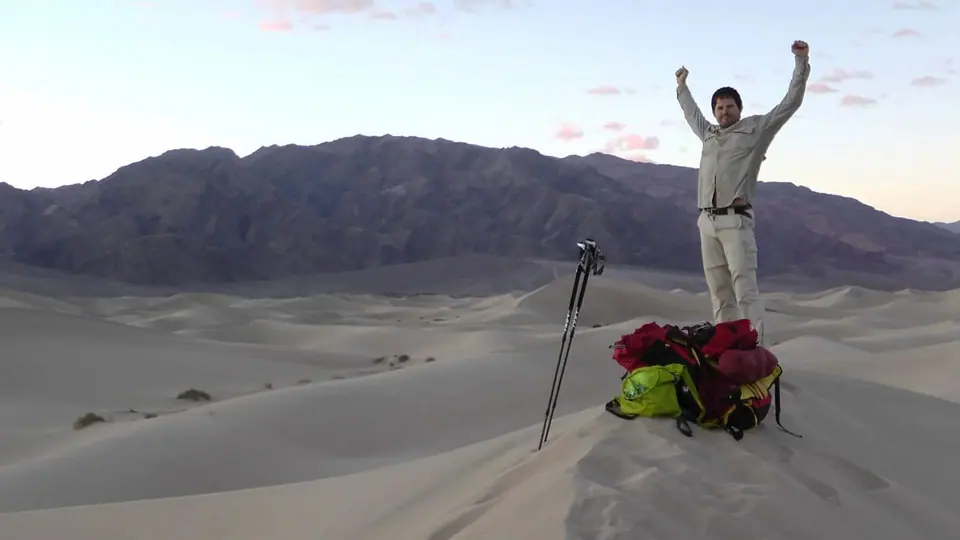 A man raising both arms on top of a sand dune beside his backpack and walking pole