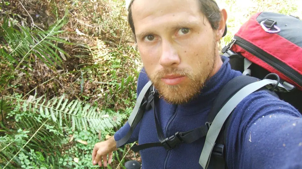 A bearded hiker with a large backpack taking a selfie among ferns