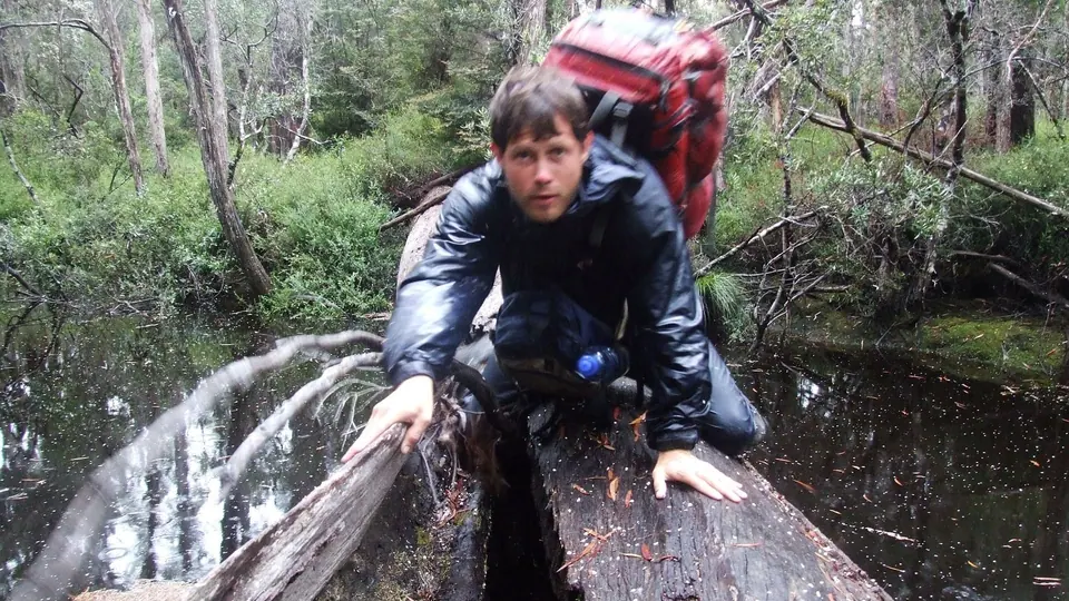 A hiker crawling along a fallen tree trunk over a stream in dense forest