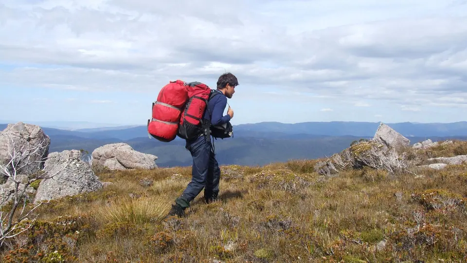 A hiker with a large red pack walking across a rocky ridge above a wide valley