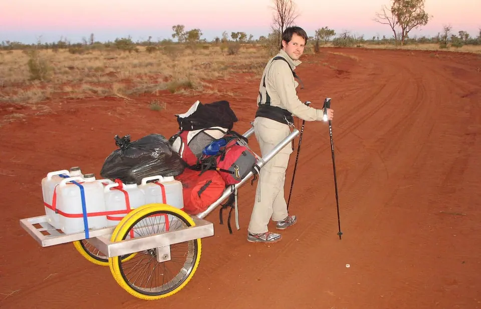 A walker pulling a cart loaded with water containers along a red desert track at dusk