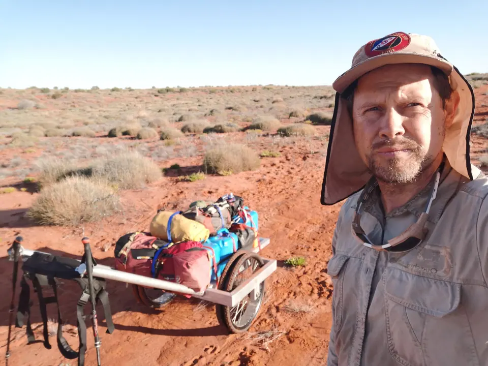 A man in a sun hat with a loaded expedition cart behind him on desert plains