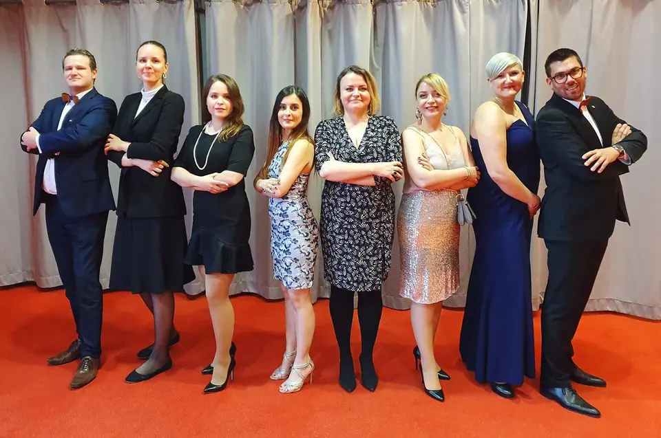 Seven people in formal wear standing in a row in front of curtains