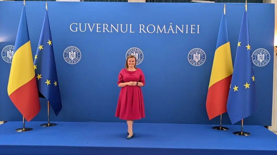 A woman in a red dress standing in front of Romanian and EU flags and a Romanian government backdrop