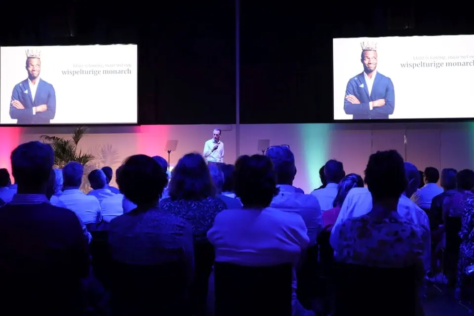 A speaker on stage in front of a seated audience, with his portrait on two screens