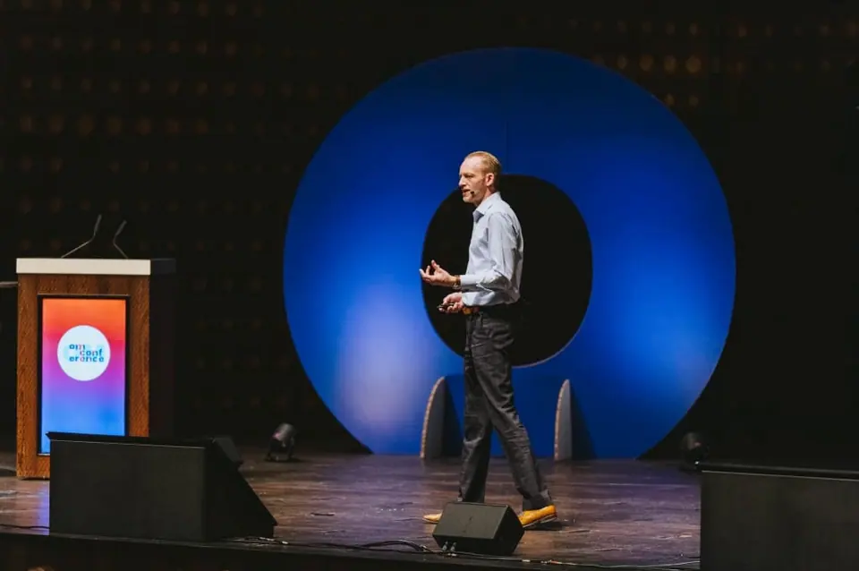 Stef Verbeeck speaking on a stage in front of a large blue ring and a lectern