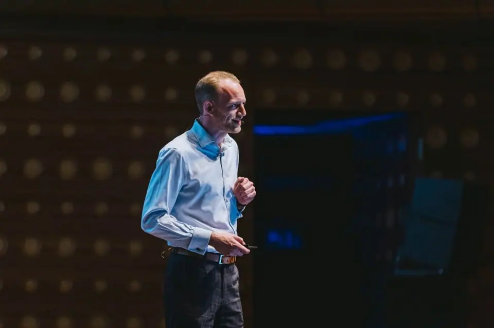 Stef Verbeeck in a light blue shirt speaking on a dark stage, holding a clicker
