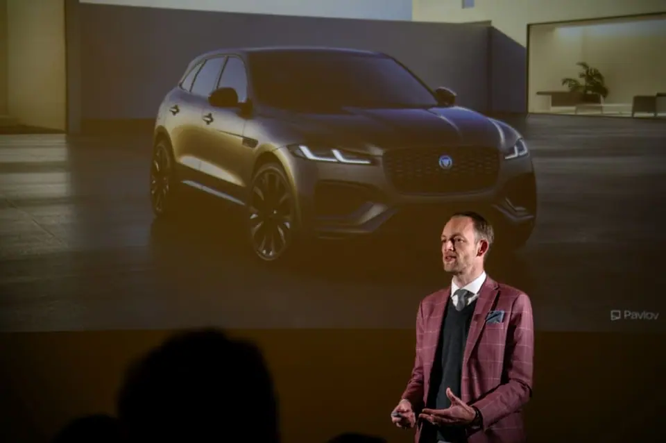 Stef Verbeeck in a burgundy blazer speaking on stage in front of a slide showing a dark car