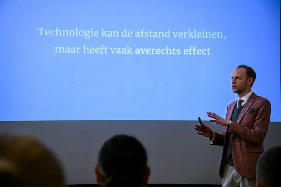 Stef Verbeeck gesturing on stage next to a slide in Dutch about technology and distance